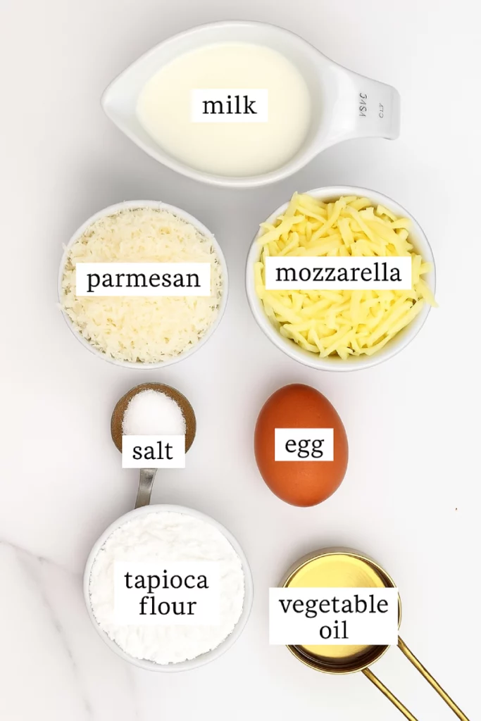 A flat lay of ingredients for Brazilian Cheese Bread, including tapioca flour, grated cheese, eggs, milk, and butter.